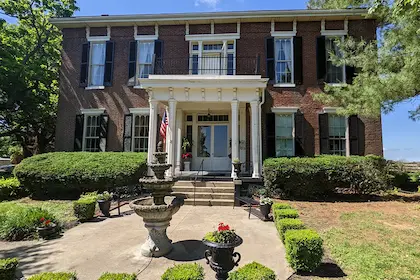 A brick, two-story historic house with a front porch, white columns, a stone fountain, and manicured landscaping.