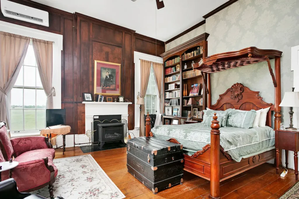 A traditional bedroom features a wood-paneled wall, fireplace, four-poster bed, bookshelf, and a red armchair.