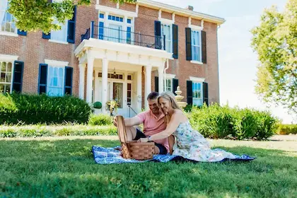 A couple has a picnic on a blue blanket in front of a large, two-story historic brick house.