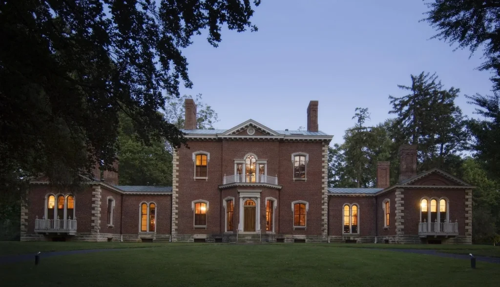 A symmetrical brick mansion with illuminated windows at dusk, surrounded by trees and a grassy lawn.