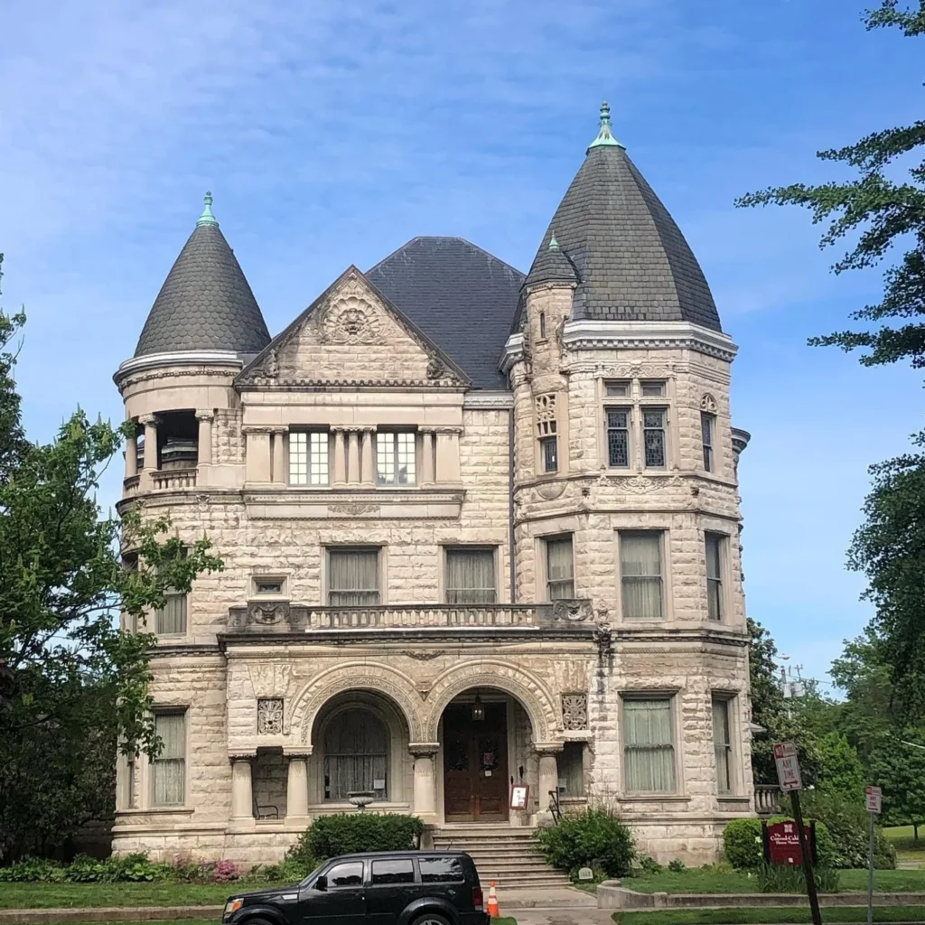 A historic, light-colored stone Victorian mansion features two prominent conical turrets under a clear blue sky.