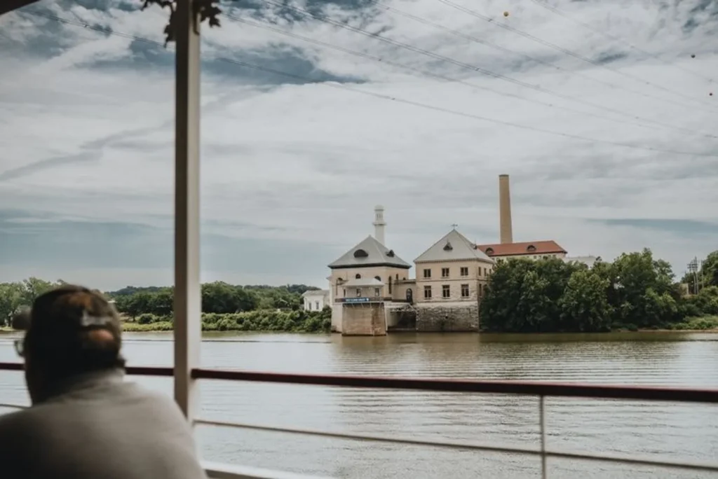 A view from a boat deck across a river toward a historic stone building complex on the far bank.