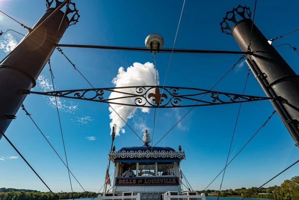View of the Belle of Louisville steamboat's cabin from the deck, showing smokestacks and white steam against blue sky.