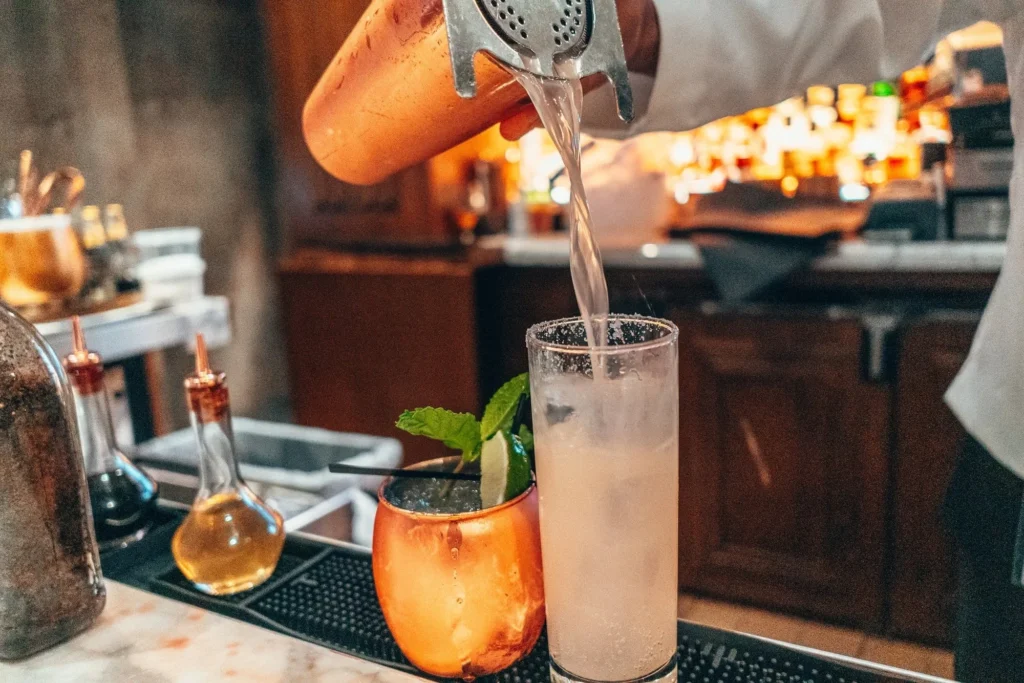 A bartender pours a cocktail from a shaker into a tall glass at a bar with other drinks in the background.