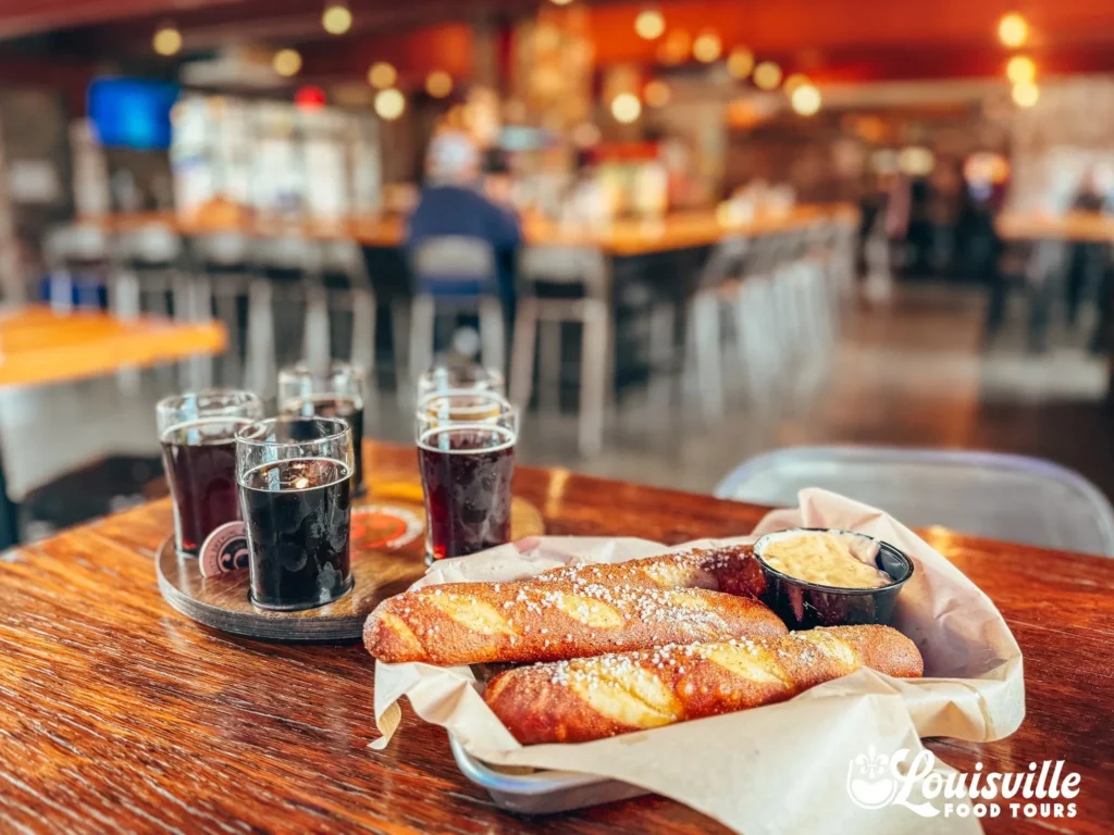 A basket of salted pretzel sticks with dipping sauce and a beer flight on a wooden table in a blurry restaurant.