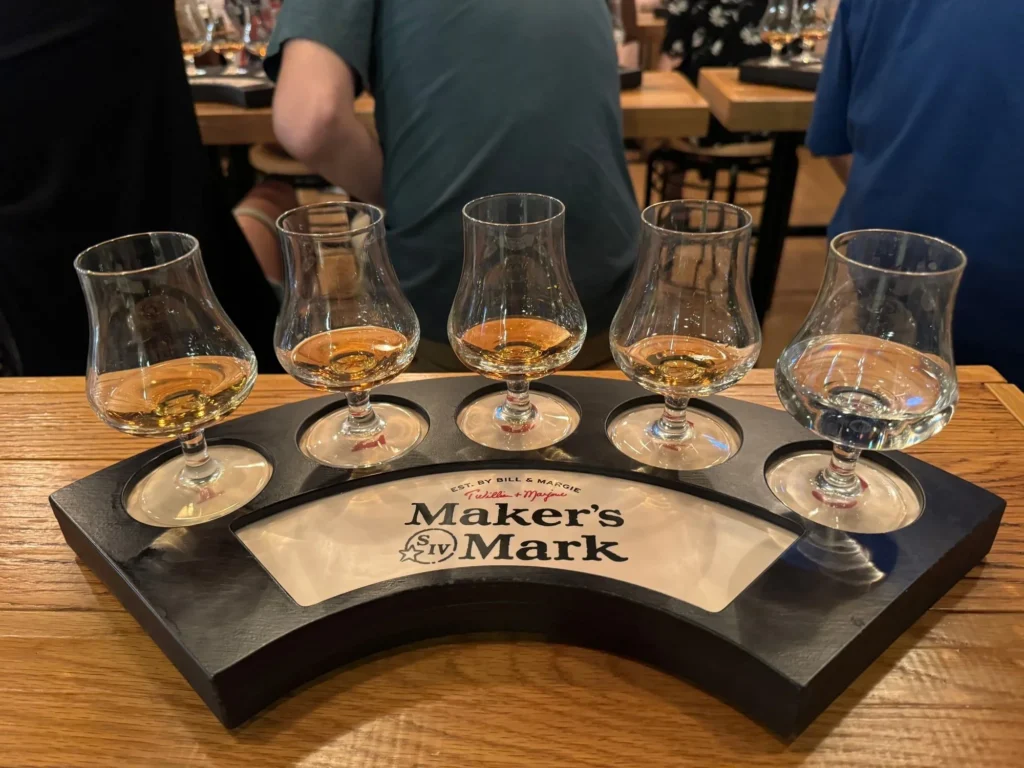 A curved Maker's Mark wooden flight tray holds five tasting glasses filled with various amber-colored spirits.
