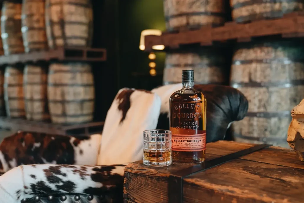A bottle of Bulleit Bourbon and a glass of whiskey sit on a wooden table in front of a chair and stacked barrels.