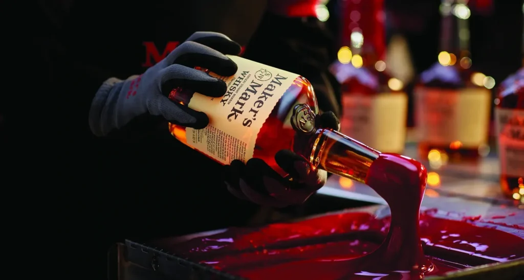 A person in gloves dips the neck of a Maker's Mark whiskey bottle into red wax.
