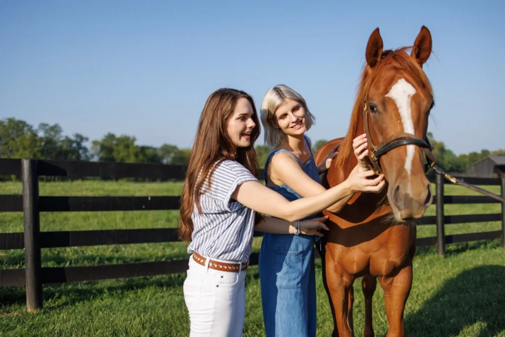 Two women stand in a grassy field by a black fence, smiling as they touch a brown horse.