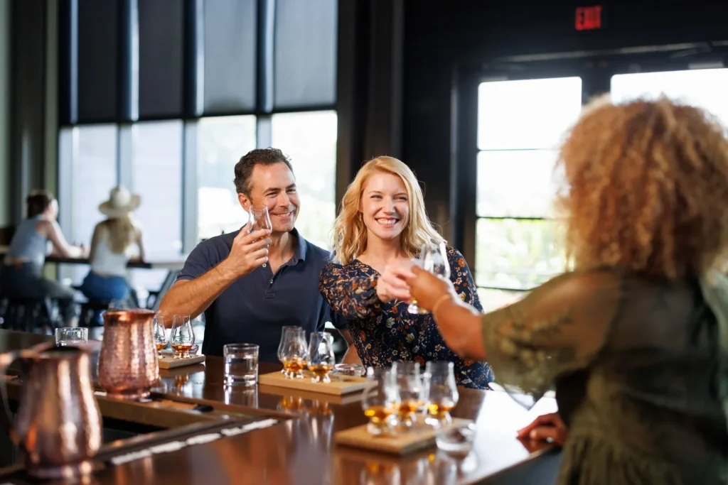 Three people raise glasses for a toast at a bar counter with tasting flights of liquor visible on the wood surface.