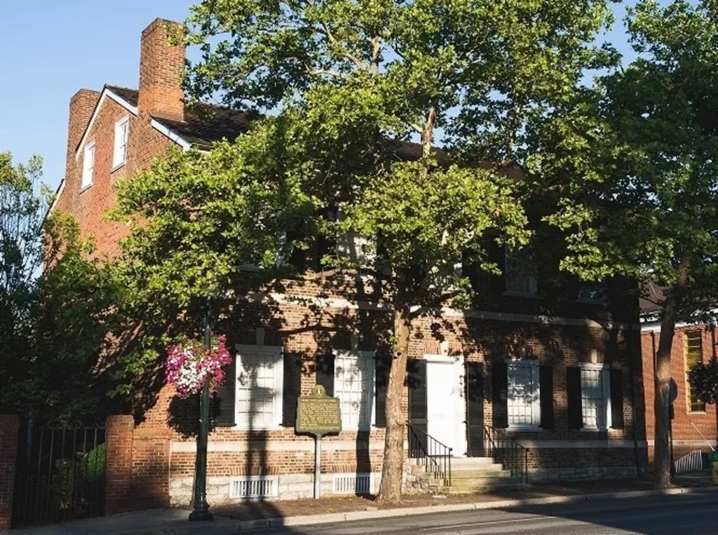 A two-story historic brick building with white shutters is partially obscured by trees and a street-side marker.