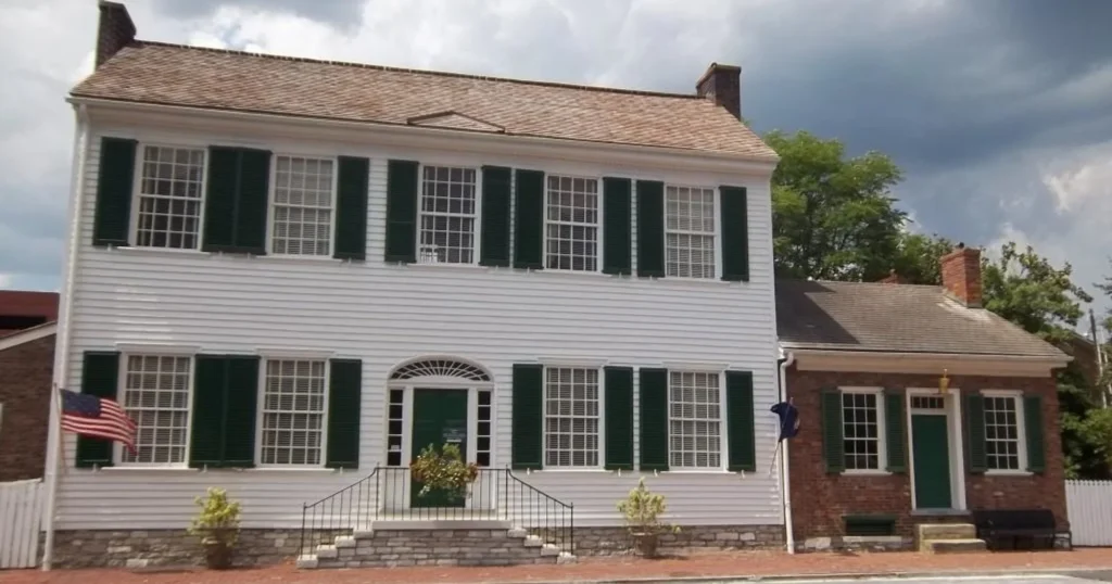 A white, two-story historic colonial house with dark green shutters and an attached brick section to the right.