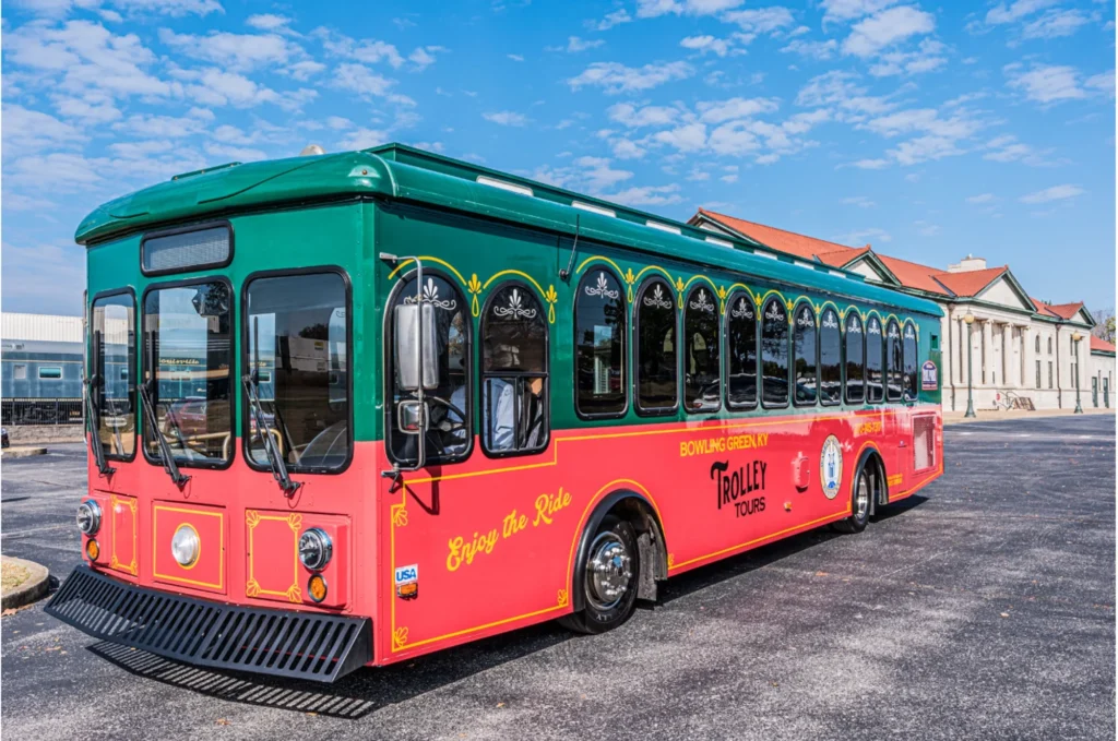 A red and green trolley bus parked in a lot in front of a building under a blue, partly cloudy sky.