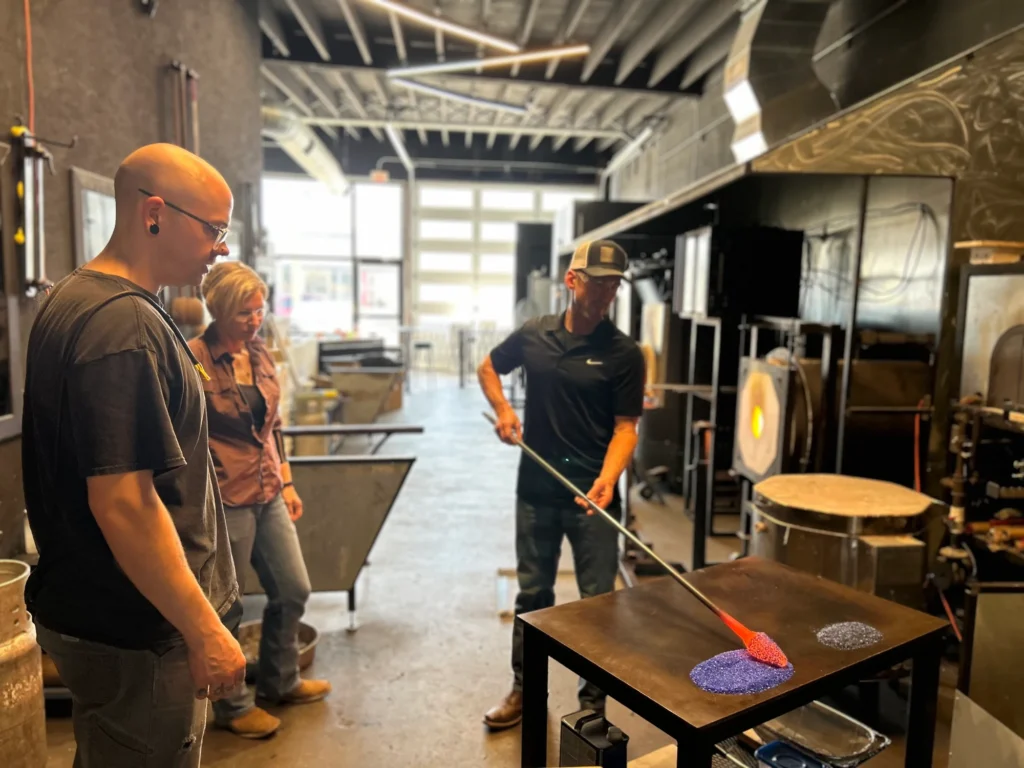 A glassblower works on a molten piece at a table while two people observe in an industrial workshop.