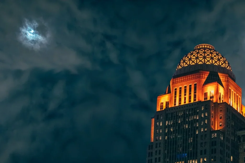 The illuminated top of a building stands against a cloudy night sky with a glowing moon.
