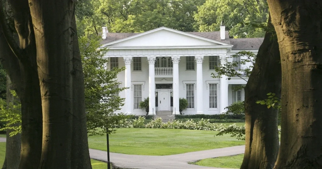 A white colonial-style mansion with tall columns is framed by trees, looking out over a green lawn and a pathway.