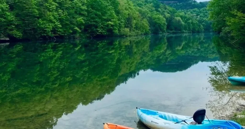 Blue and orange kayaks rest on a shore by a calm, green river reflecting lush forest trees.
