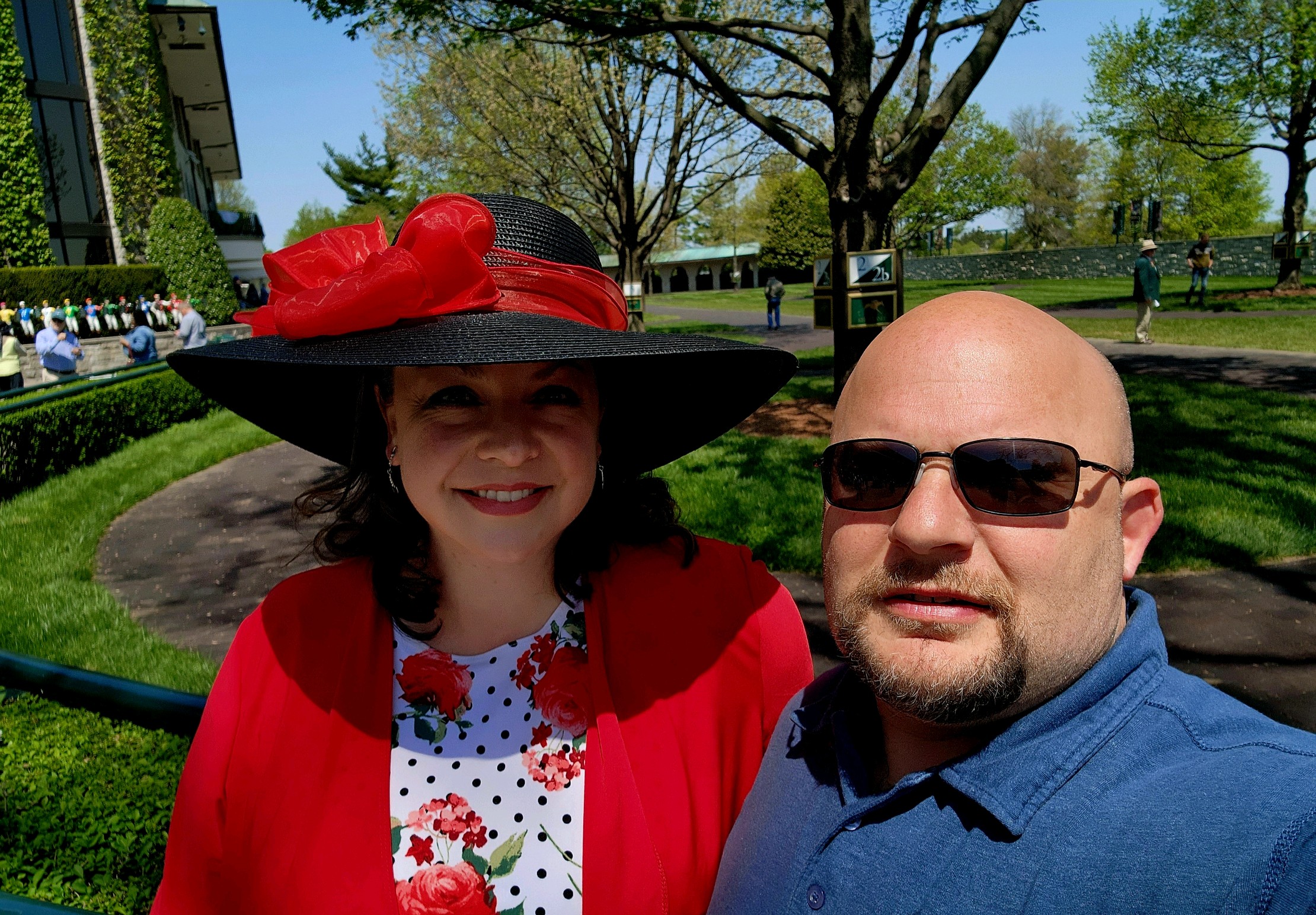 A woman in a red hat and a man in sunglasses pose for a selfie outdoors on a sunny day.