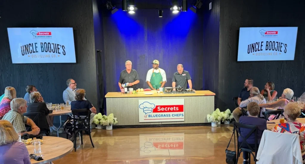 Three men stand behind a demo table on a stage in front of a seated audience, with "Uncle Boojie's" screens displayed.